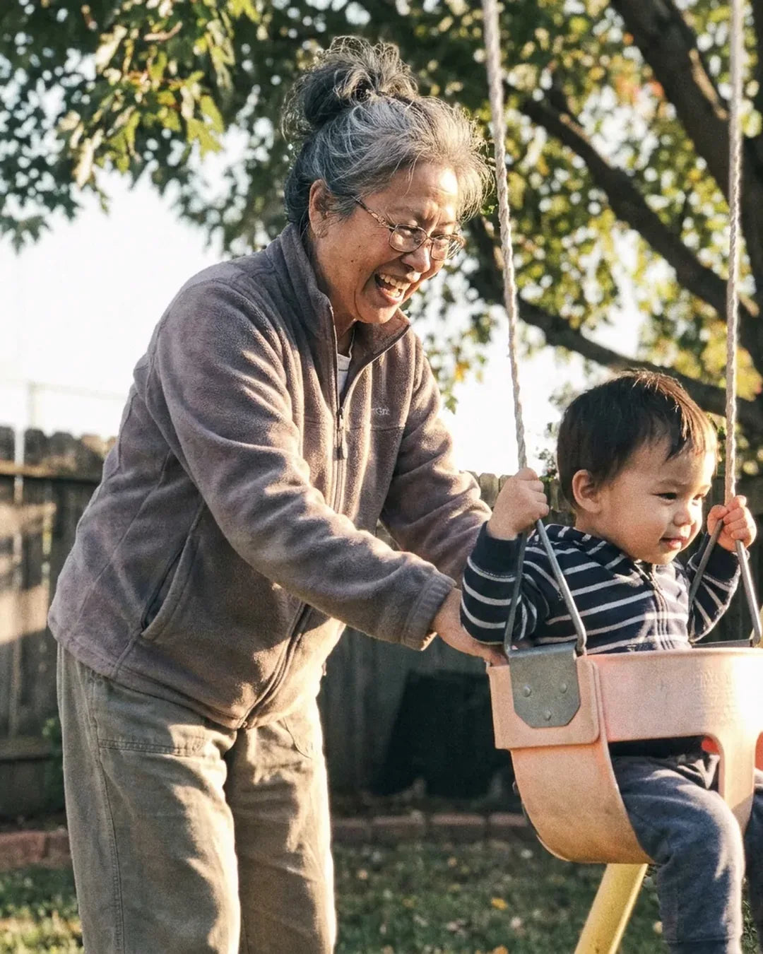 A woman in her sixties laughing as she pushes a small grandchild on a backyard swing at golden hour