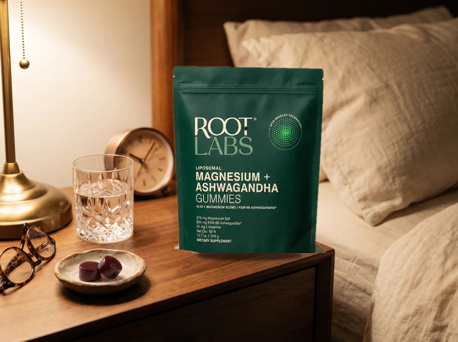 AshwaMag pouch on a warm oak nightstand: two berry gummies in a small ceramic dish, cut-glass water tumbler, folded reading glasses, hardcover book, and a warm brass lamp - the nightly ritual that women 45+ actually keep